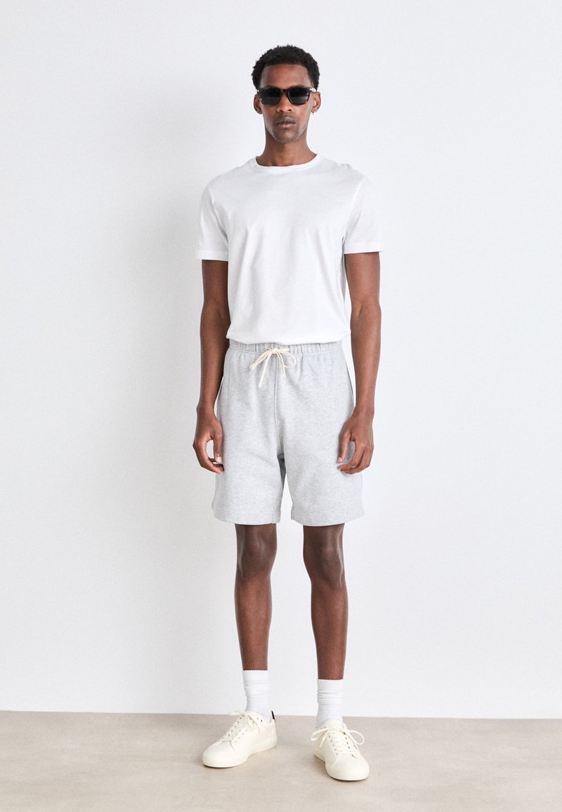White t-shirt, gray shorts with drawstring, and beige sneakers. Model wears black sunglasses, stands against a plain backdrop.