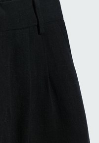Pantaloni neri su misura in primo piano, caratterizzati da una vita piatta, una superficie liscia e dettagli di pieghe precise in vita.