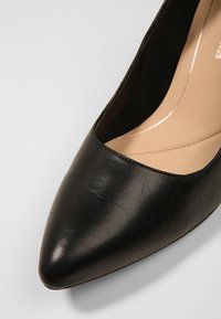 Chaussure à talon haut en cuir noir avec un bout pointu et une texture lisse. L'intérieur présente une doublure beige avec des détails de couture subtils.