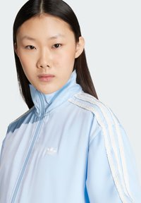 Svetlomodrá bunda na zips s vysokým golierom, bielymi pruhmi a vyšitým logom na hrudi, vyrobená z hladkej, ľahkej tkaniny.