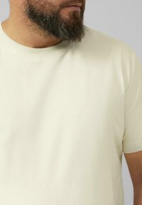 Jasno beżowy t-shirt wykonany z bawełny, z okrągłym dekoltem, krótkimi rękawami i gładką fakturą. Ubranie ma subtelne szwy i minimalne wykończenia.