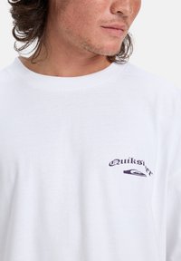 Hvit bomulls T-skjorte med en mørk lilla "Quiksilver" logo på venstre brystområde. Rund hals og avslappet passform. Myk stofftekstur.