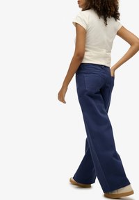 Camisa corta de manga corta en beige claro combinada con vaqueros de pierna ancha de color azul oscuro, con bolsillos traseros y un ajuste holgado, llevada con zapatos claros.