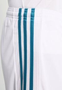 Valkoiset urheilushortsit, joissa on teal-väriset pystyraidat sivuilla. Shortsit ovat valmistettu tekstuurista kankaasta ja niissä on joustava vyötärönauha mukavuutta varten.