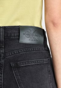 Close-up of black Calvin Klein jeans waist label worn with a pale yellow shirt and visible arm.