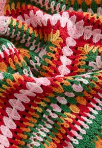 Manta de punto multicolor que presenta hilo rojo, verde, naranja, rosa y blanco en un patrón texturizado con bordes ondulados y puntadas abiertas.