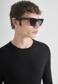Occhiali da sole neri rettangolari con lenti scure, abbinati a un maglione nero a coste con scollatura rotonda e motivo testurizzato.