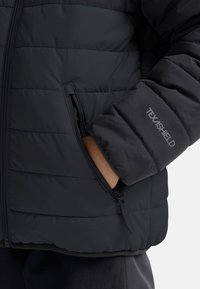 Chaqueta acolchada negra con un bolsillo con cremallera y la marca TEXSHIELD en el puño, con una textura mate y suave.
