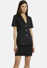 Donker blazer met korte mouwen, hoofdkraag, drie grote knopen en twee voorzakken. Gecombineerd met een bijpassende getailleerde rok.