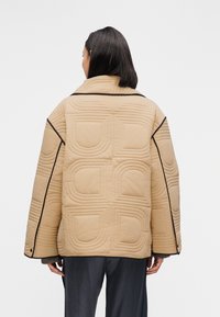 Μπεζ κουμπωτό μπουφάν με ανάγλυφα γεωμετρικά μοτίβα, μαύρο περίγραμμα και δομημένο γιακά. Διαθέτει φαρδιά γραμμή και υφή υφάσματος.