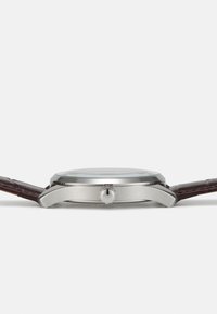 Reloj de acero inoxidable plateado con caja redonda, correa de cuero marrón y una corona con un logotipo. Cubierta de cristal suave sin marcas visibles.