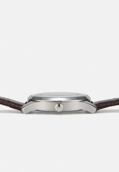 Montre en acier inoxydable avec un boîtier circulaire, un bracelet en cuir marron et un détail de couronne. Elle possède un cadran en verre transparent et un design minimaliste.