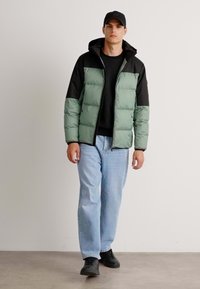 Pufferjack met een groen en zwart kleurenschema, voorzien van een ritssluiting aan de voorkant, capuchon, geborduurde textuur en gecombineerd met lichte jeans en zwarte schoenen.