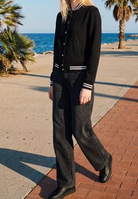 Jeune personne marchant sur une promenade au bord de la mer portant une veste universitaire noire, un jean large foncé et des baskets noires. Palmiers à proximité.