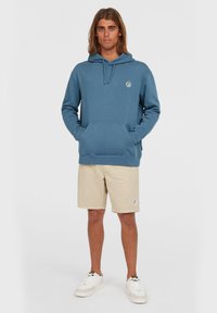 Blue hooded sweatshirt with a front pocket, featuring a circular logo. Paired with beige shorts and white sneakers. Smooth texture.