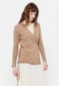 Light brown knit wrap sweater with long sleeves, deep V-neck, and side tie detail. Paired with a ribbed, creamy white skirt.