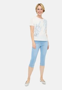 Weißes T-Shirt mit blauem Blumenmuster, kombiniert mit hellblauen, verkürzten Jeans. Das Model trägt silberne Ballerinas, steht und lächelt.