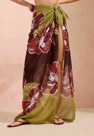 Langer grüner und weinroter Blumensarong, an der Taille gebunden, getragen mit weinroten Sandalen, zeigt ein Bein durch einen Frontschlitz.