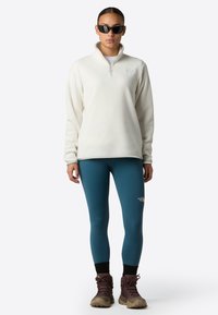 Crème fleece pullover met een kwart-rits, blauwe leggings en bruine wandelschoenen. Zwarte zonnebril maakt de outfit compleet. Logo zichtbaar op de top en leggings.