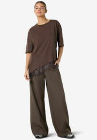 Brūna, vaļīga T-krekls ar īsām piedurknēm un mežģīņu apdari apakšmalā. Saskaņots ar brūnām plaša piegriezuma biksēm un sudraba sporta apaviem.