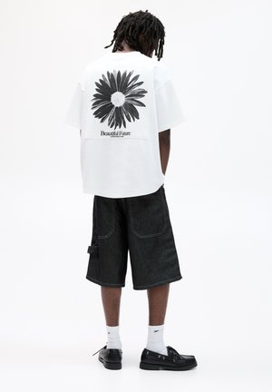 Hvid oversized t-shirt med et sort tusindfryd grafik og teksten "Beautiful Future"; kombineret med mørk denim shorts og sorte sko.