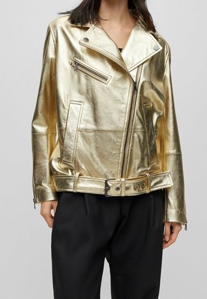 Kvinde iført en metallisk gylden bikerjakke med lynlåslommer og bælte i taljen, kombineret med løse sorte bukser mod en neutral baggrund.
