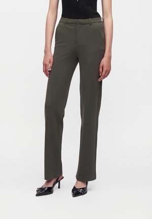 Femme portant un pantalon droit vert olive, un top noir sans manches et des chaussures à talons aiguilles noirs et pointus avec des boucles décoratives.
