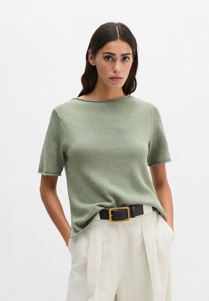 Woman wearing a light green short-sleeve knit top tucked into high-waisted white pants with a black belt featuring a large gold buckle.