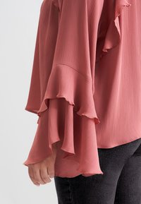 Blusa a maniche lunghe in rosa ruggine con trama a coste, caratterizzata da maniche svasate con bordi ondulati e orlo arrotondato. Abbinata a jeans scuri.