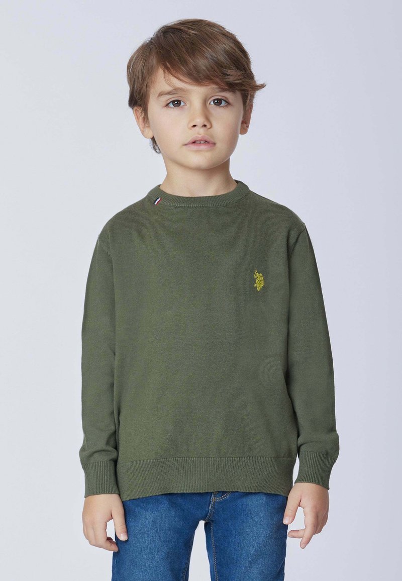 Maglione verde a maglia con collo rotondo, dotato di un piccolo logo giallo e di un accento a contrasto vicino al colletto. Abbinato a jeans blu.