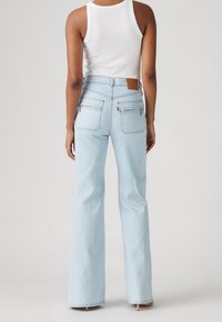 Woman wearing light blue high-waisted jeans with back zipper pockets and a white sleeveless ribbed top, standing against a plain background.