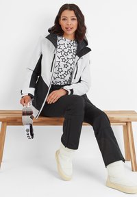 Snow jacket in white and black with a hood, featuring zippered pockets. Black pants and glossy white insulated boots. Holding ski goggles.