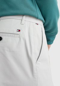 Persona con pantaloni beige e un piccolo logo rosso, bianco e blu vicino alla tasca, e una felpa teal, mostrata dalla vita in giù in vista laterale.
