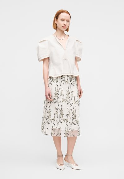 Μπλούζα σε χρώμα κρέμα με φουσκωτά μανίκια και φερμουάρ μπροστά, συνδυασμένη με μίντι φούστα με φλοράλ τύπωμα, που έχει αέρινη, διάφανη υφή. Άσπρα τακούνια.