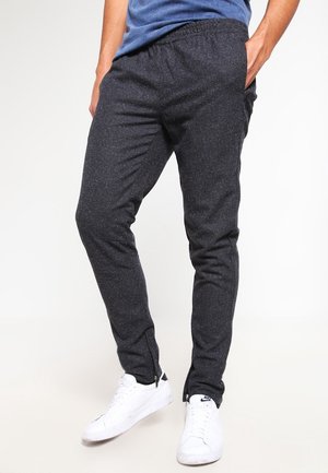 Man draagt donkergrijze taps toelopende joggerbroek en witte Nike sneakers, staat met één hand in de zak tegen een witte achtergrond.