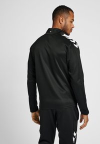 Veste de sport noire à manches longues, col haut, avec des accents en chevron blancs sur les épaules. Texture lisse, design ajusté.
