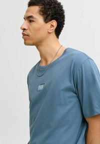 Camiseta azul de algodón con mangas cortas, que presenta un pequeño logo rectangular en el área del pecho izquierdo y un cuello redondo.