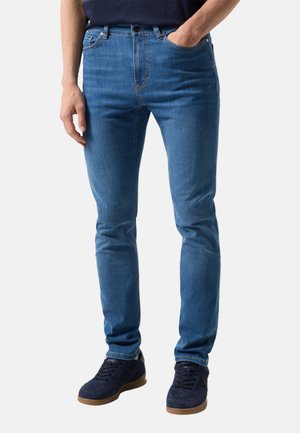 Mężczyzna noszący dopasowane niebieskie dżinsy i ciemnoniebieskie trampki, z jedną ręką w kieszeni, stojący na jednolitym tle.