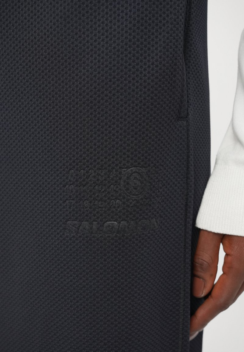 Tissu texturé noir avec un motif en relief, présentant une marque SALOMON en relief et des détails sur le produit. Placé à la main sur le côté.