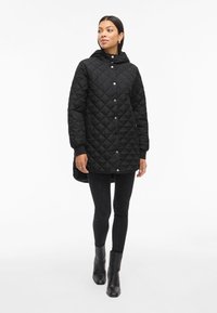 Črna prešitka jakna s kapuco, sprednjim zapiranjem na pritiskače in rebrastimi manšeti. Oblečena čez črne skinny hlače in gležnjarje.