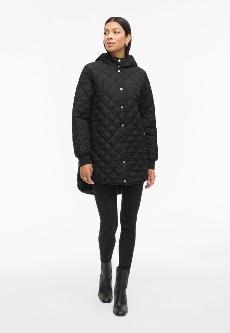 Črna prešitka jakna s kapuco, sprednjim zapiranjem na pritiskače in rebrastimi manšeti. Oblečena čez črne skinny hlače in gležnjarje.