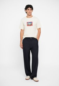 Beige bomulds t-shirt med et bjergmotiv omkranset af rødt, parret med mørke smalle stribede bukser og lyse sneakers.