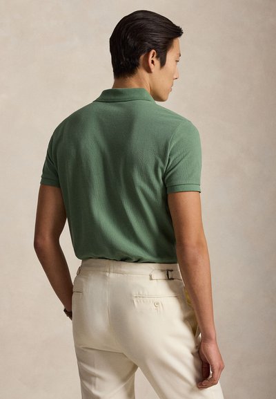 Vyras stovintis nugara, dėvintis žalią trumpomis rankovėmis polo marškinėlį ir smėlio spalvos aukšto liemens kelnes ant vientisos fono.