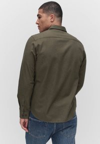 Olijfgroene lange mouw shirt met een puntige kraag en knopenmanchetten, gemaakt van een gladde, gestructureerde stof. Draagt met een blauwe spijkerbroek.