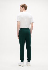 Pantaloni sportivi di un verde scuro in misto cotone, con vita regolabile tramite cordoncino e polsini a coste, abbinati a una t-shirt bianca semplice.