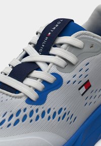 Λευκό αθλητικό παπούτσι με μπλε λεπτομέρειες, επάνω μέρος από δίχτυ με οπές αερισμού και μπλε λουρί που φέρει το όνομα της μάρκας και το λογότυπο.