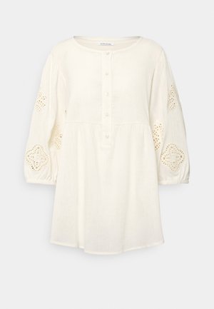 Hvid tunika med lange ærmer, rund halsudskæring, knaplukning foran og dekorative blonderudskæringer på puffede ærmer.