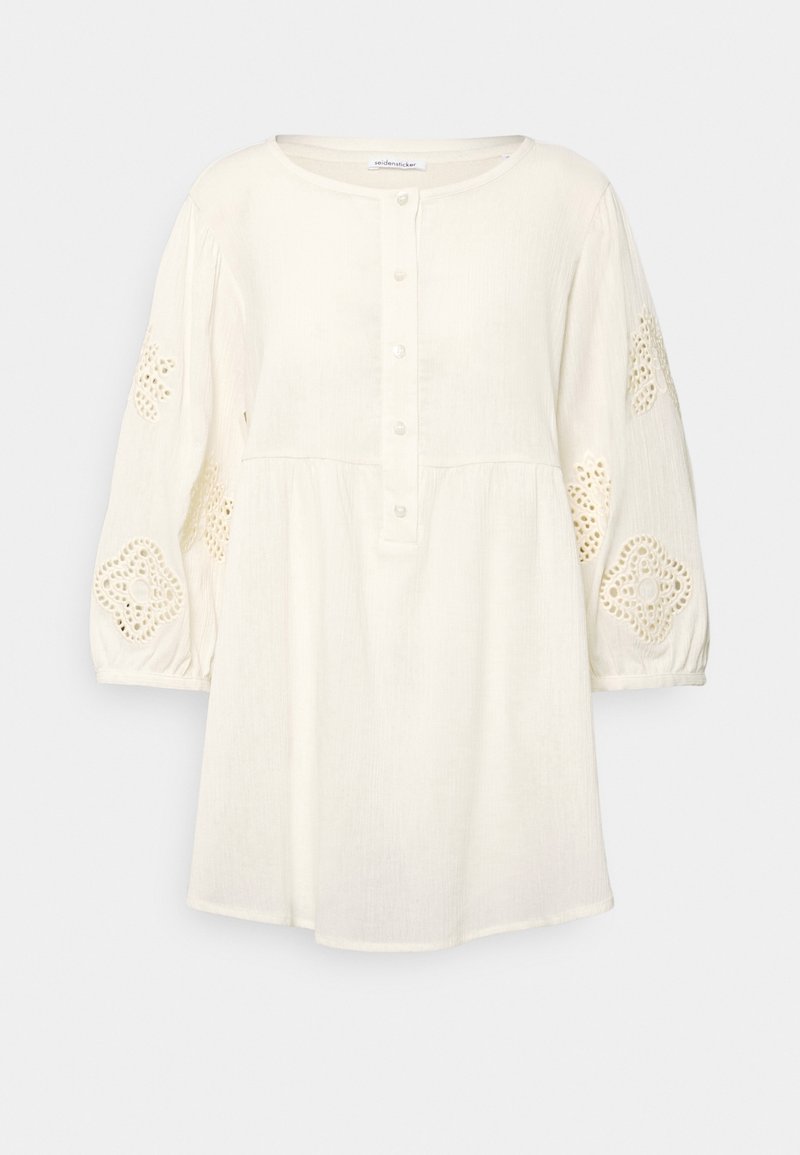 Tunique blanche à manches longues avec encolure ronde, patte de boutonnage à l'avant et découpes décoratives en dentelle sur les manches bouffantes.