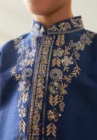 Indumento blu navy ricamato con intricati motivi floreali dorati e paillettes. Presenta un colletto a fascia e un design della scollatura dettagliato.