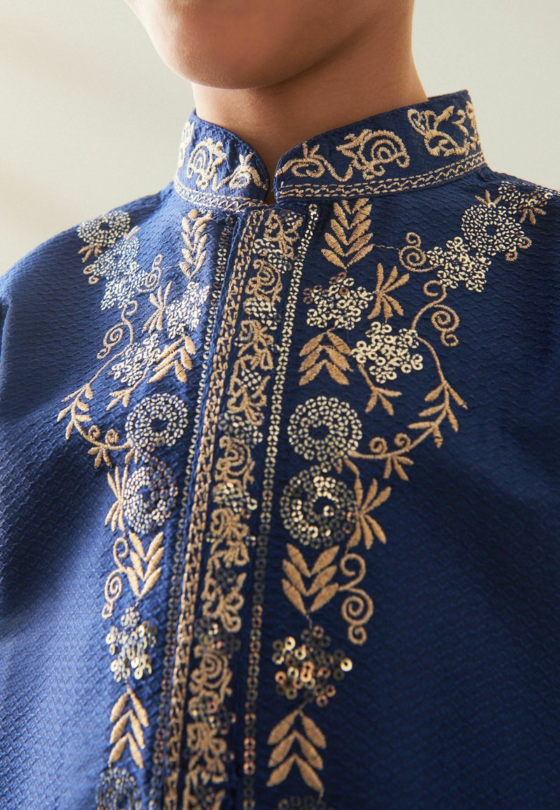 Indumento blu navy ricamato con intricati motivi floreali dorati e paillettes. Presenta un colletto a fascia e un design della scollatura dettagliato.